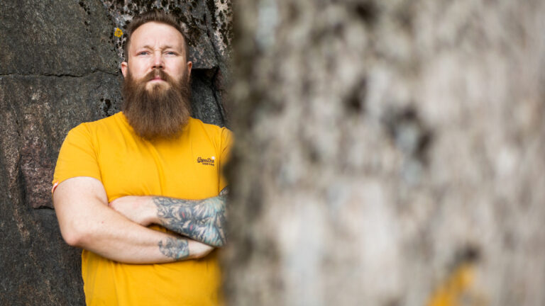 Mies katsoo kameraan kädet rinnalla, osittain puun takana.