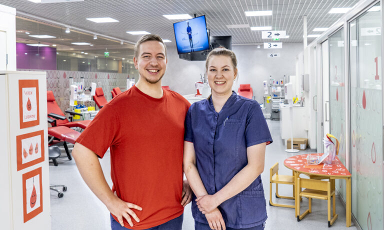 Kaksi Veripalvelun työntekijää hymyilee kameralle verenluovutuspisteessä.