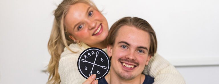 Vaaleahiuksinen nainen miehen reppuselässä, näyttävät kameralle Kantasolurekisteri-kangasmerkkiä.